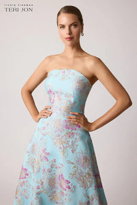 A model wearing a strapless light blue A-line gown with pink and gold floral embroidery, posed against a solid light gray background. The brand name 'Teri Jon' is visible in the top left corner.