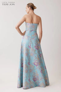A model shown from the back wearing a strapless A-line gown by Teri Jon, featuring a light blue base with a purple and gold floral jacquard pattern, against a neutral studio background.