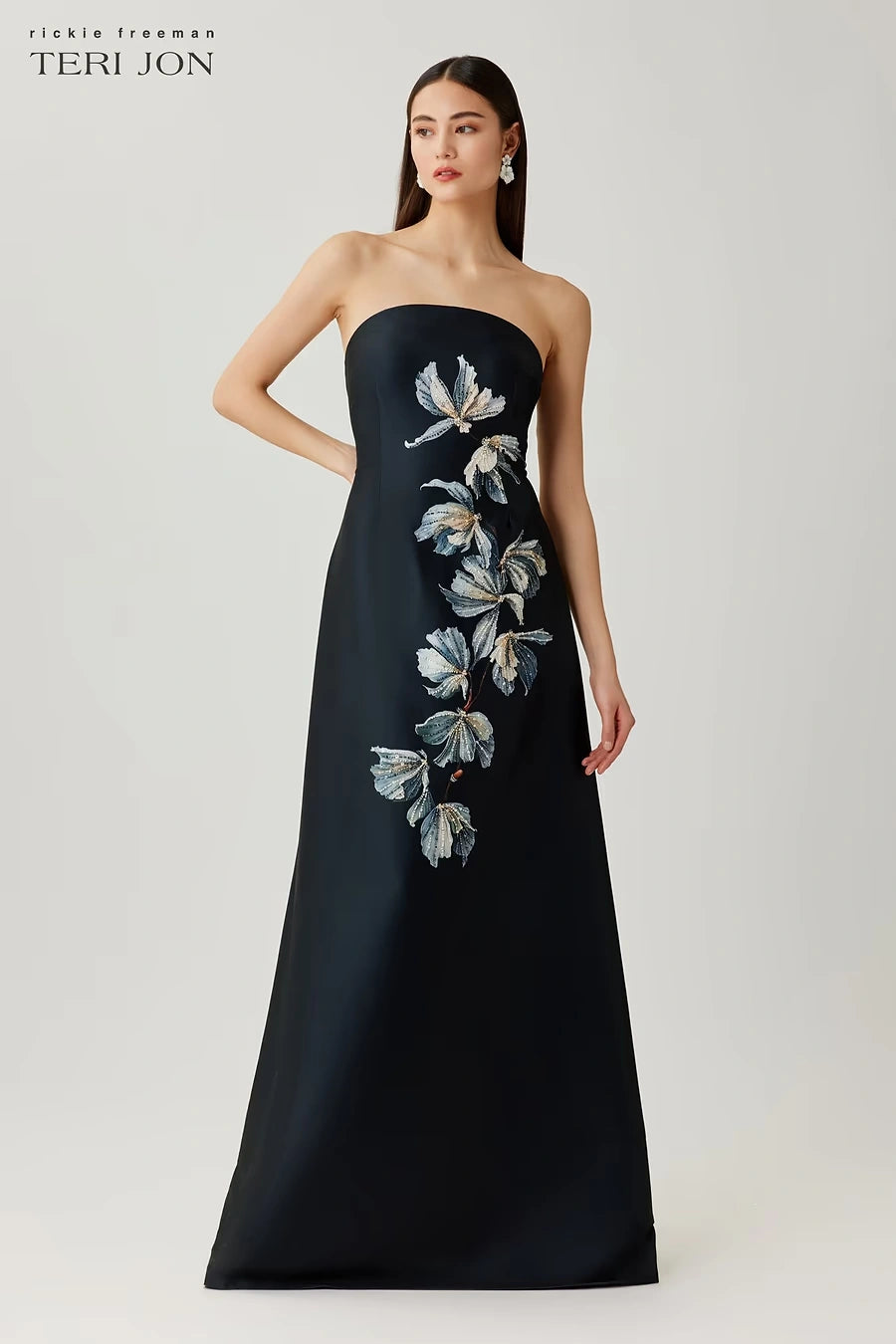 A model wears a strapless black floor-length gown by Teri Jon, featuring a vertical floral applique design in blue, white, and gray on the bodice and cascading down the left side of the skirt. The brand logo 'teri jon' is visible in the top left corner.