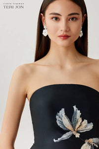 Woman wearing a black strapless dress with floral embroidery on a plain background
