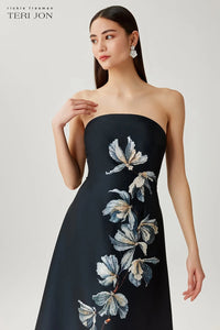 Woman wearing a black strapless dress with floral embroidery on a light gray background