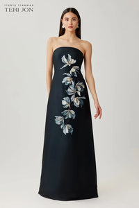 Woman wearing a black strapless gown with floral embroidery on a white background, Teri Jon brand visible.
