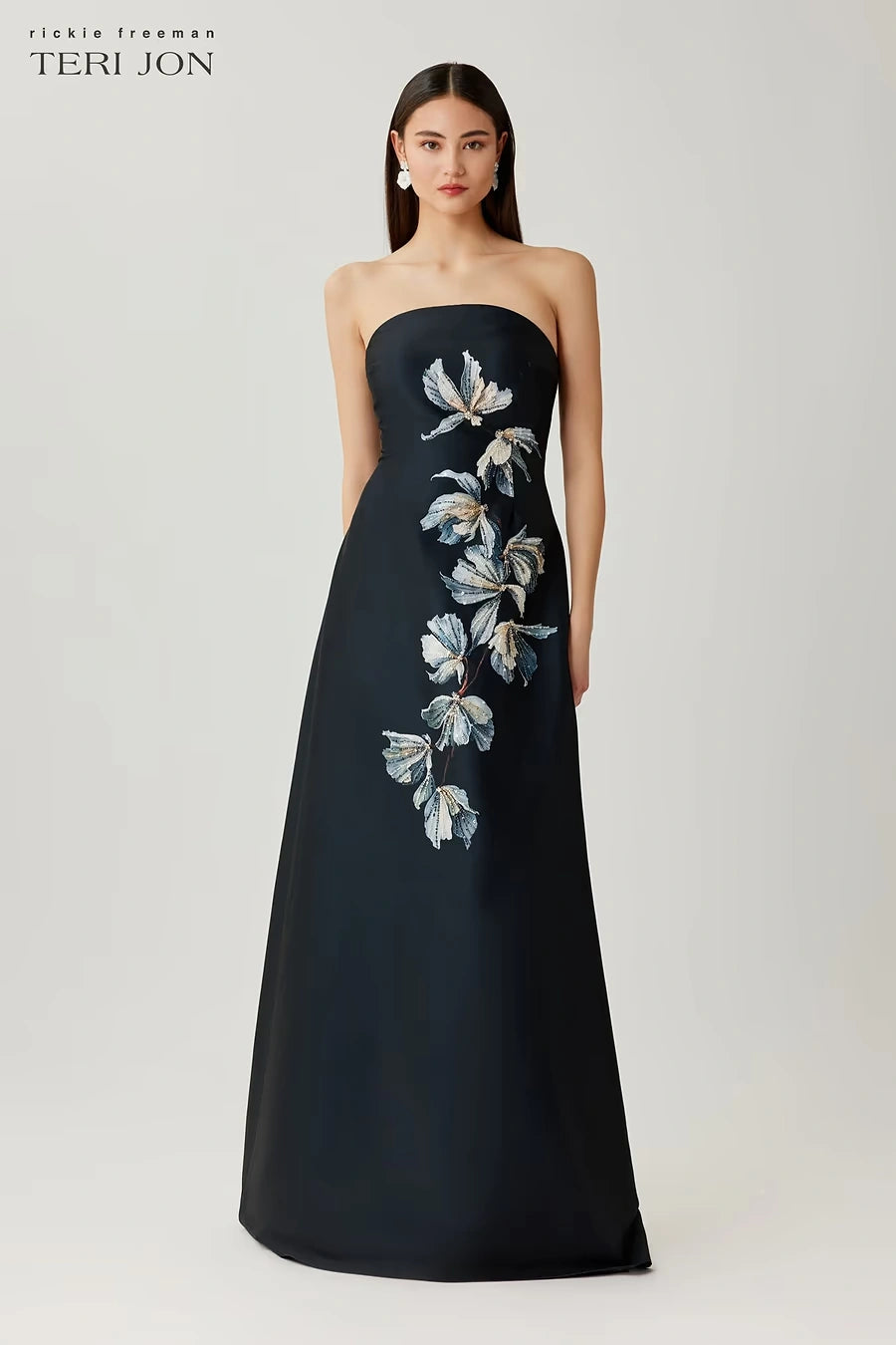 A model wears a strapless floor-length gown by Teri Jon, featuring a dark navy satin base with a vertical floral applique pattern in white, light blue, and gray on the bodice and skirt. The brand logo 'teri jon' is visible in the top left corner.