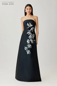 A model wears a strapless floor-length gown by Teri Jon, featuring a dark navy satin base with a vertical floral applique pattern in white, light blue, and gray on the bodice and skirt. The brand logo 'teri jon' is visible in the top left corner.