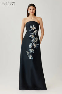 Woman wearing a black strapless gown with floral embroidery on a white background, Teri Jon brand visible.