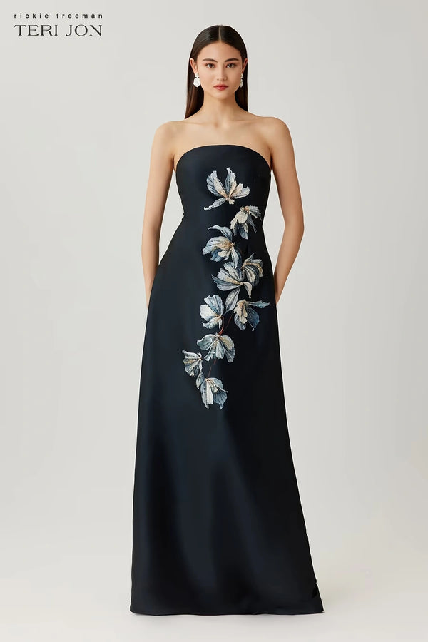 Woman wearing a black strapless gown with floral embroidery on a white background, Teri Jon brand visible.