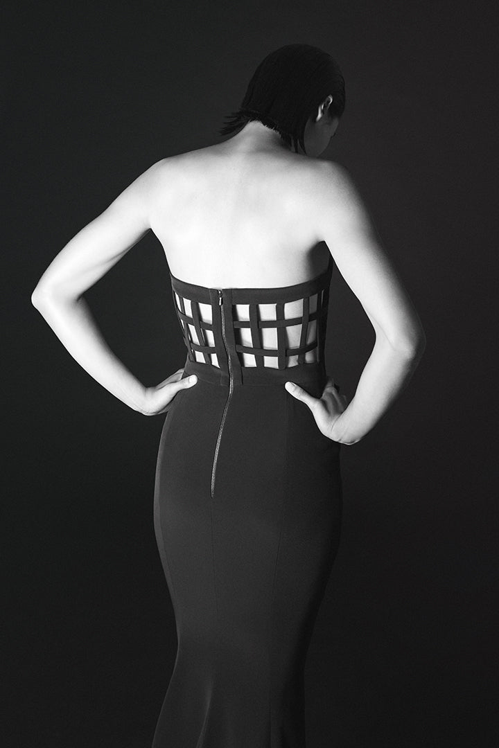 A black and white studio photograph of a model from behind, wearing a strapless black dress with a structured, grid-like cutout corset-style waist and a fitted silhouette.