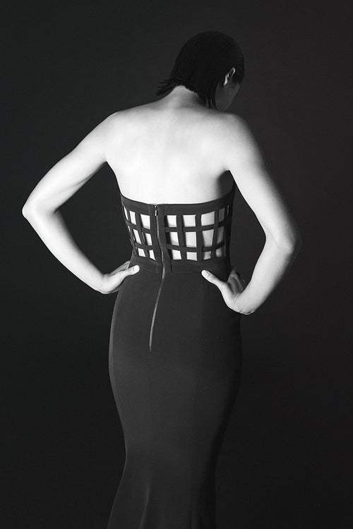 A black and white studio photograph of a model from behind, wearing a strapless black dress with a structured, grid-like cutout corset-style waist and a fitted silhouette.