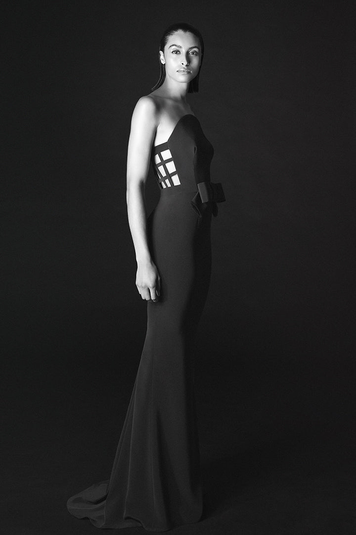 A woman modeling a floor-length black gown in a studio setting against a dark background. The dress has a strapless silhouette with a structured, geometric cage-like bodice panel and a long train.