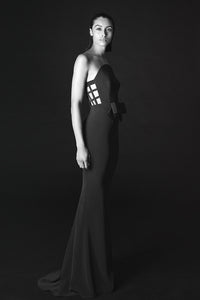 A woman modeling a floor-length black gown in a studio setting against a dark background. The dress has a strapless silhouette with a structured, geometric cage-like bodice panel and a long train.
