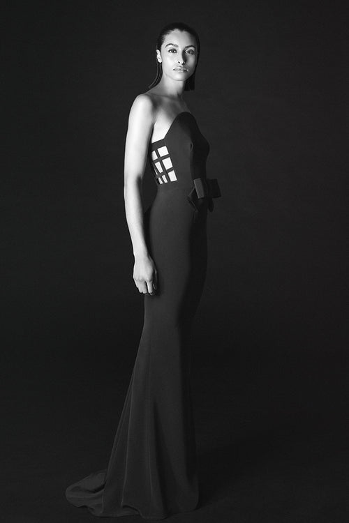 A woman modeling a floor-length black gown in a studio setting against a dark background. The dress has a strapless silhouette with a structured, geometric cage-like bodice panel and a long train.